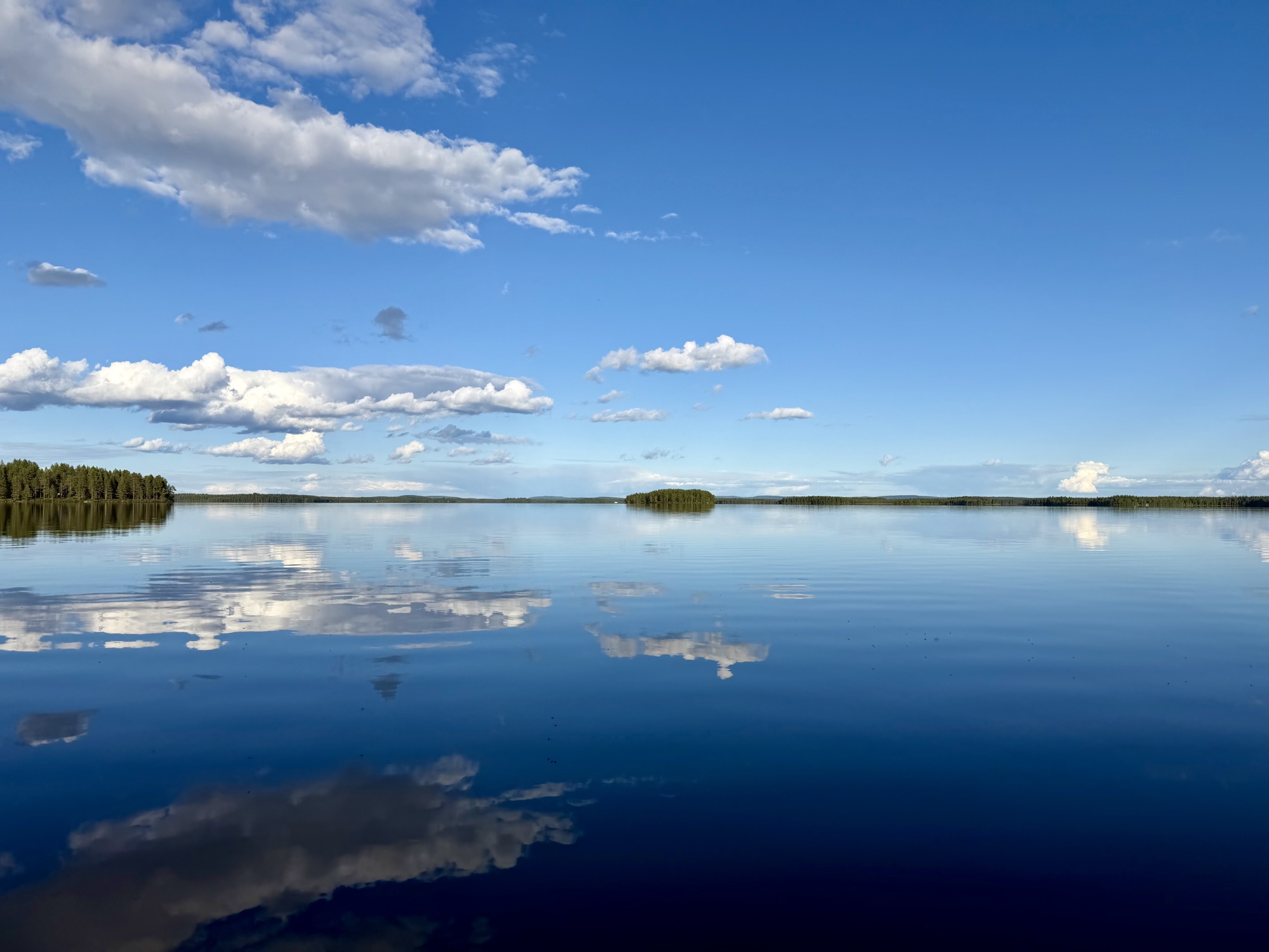 Spiegelglatter See mit Wolken, die sich im Wasser spiegeln, am Horizont eine Waldlinie