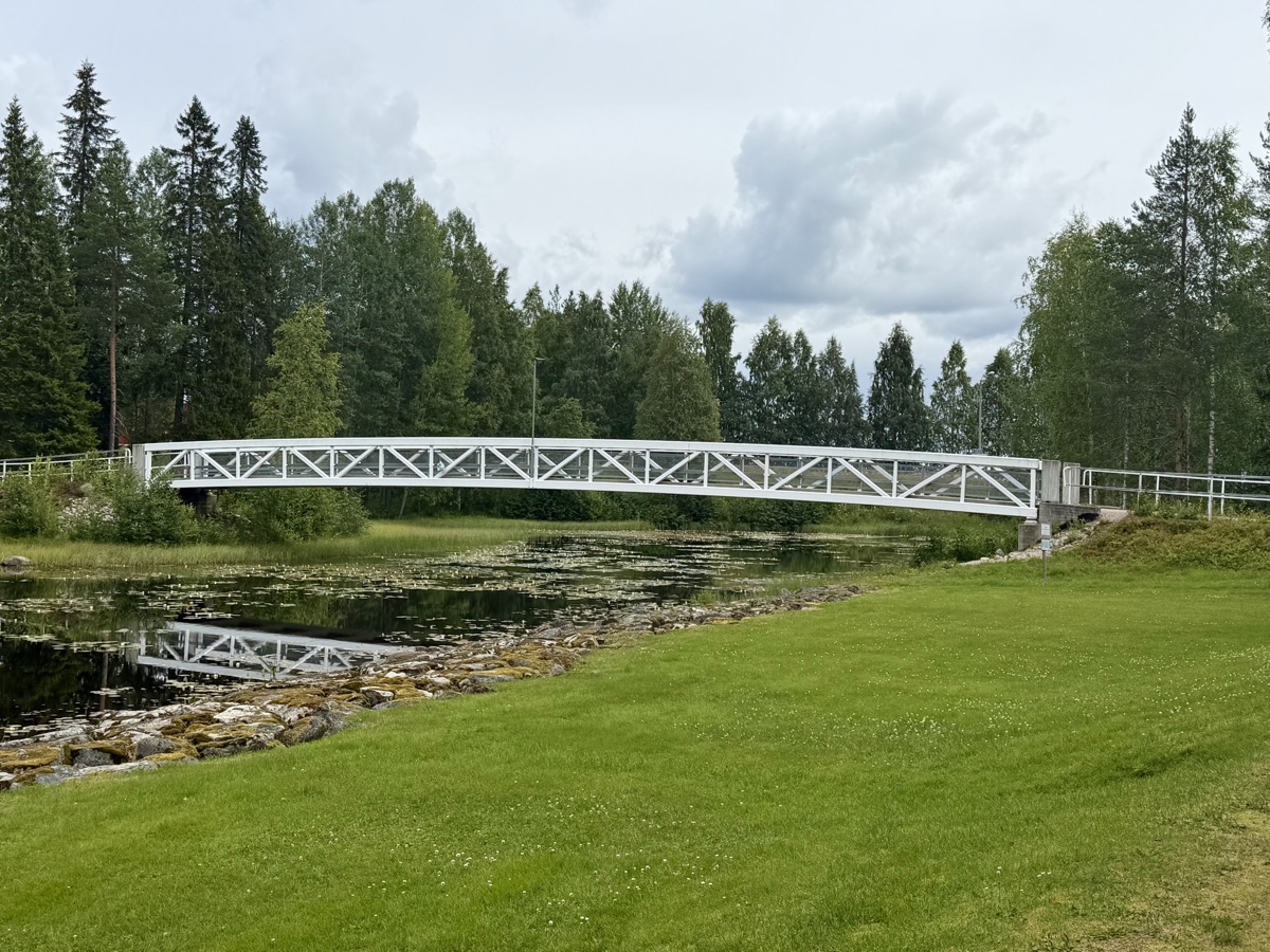 Weiße Fachwerkbrücke aus Holz spannt sich über einen stillen Flussarm, davor eine grüne Wiese