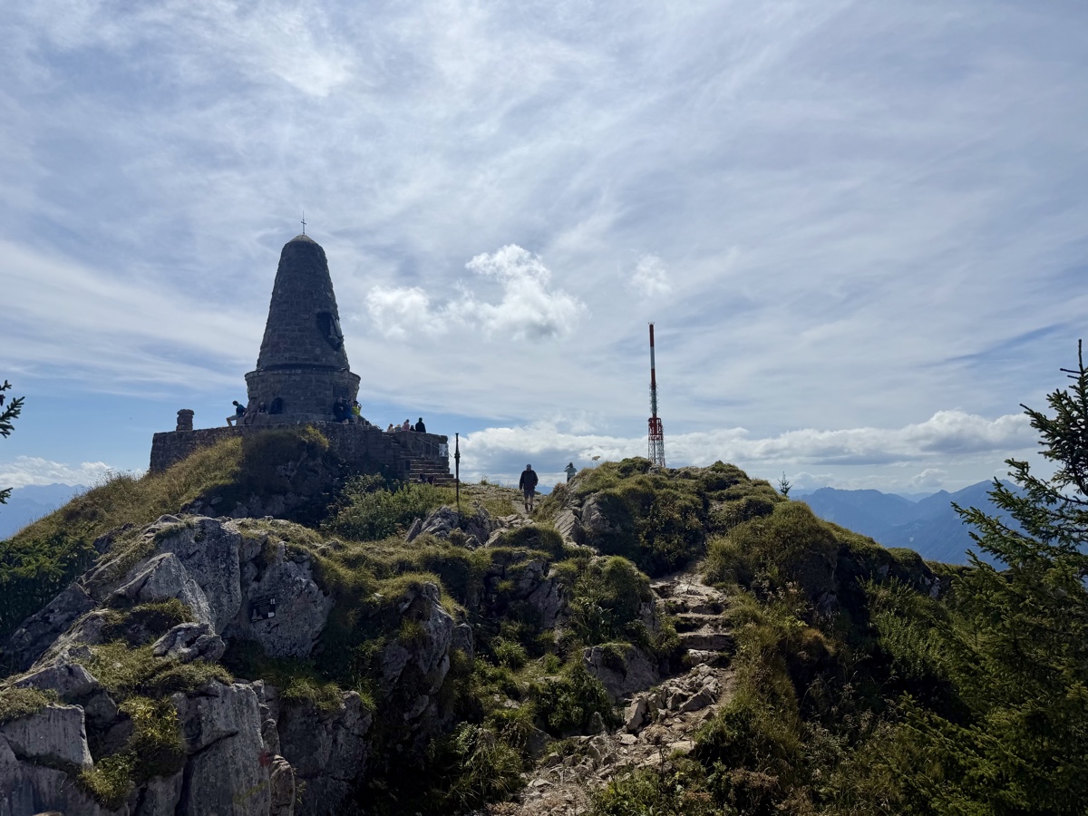 Steinernes Gipfelbauwerk mit Spitzdach auf einem Felsgrat, daneben ein Sendemast