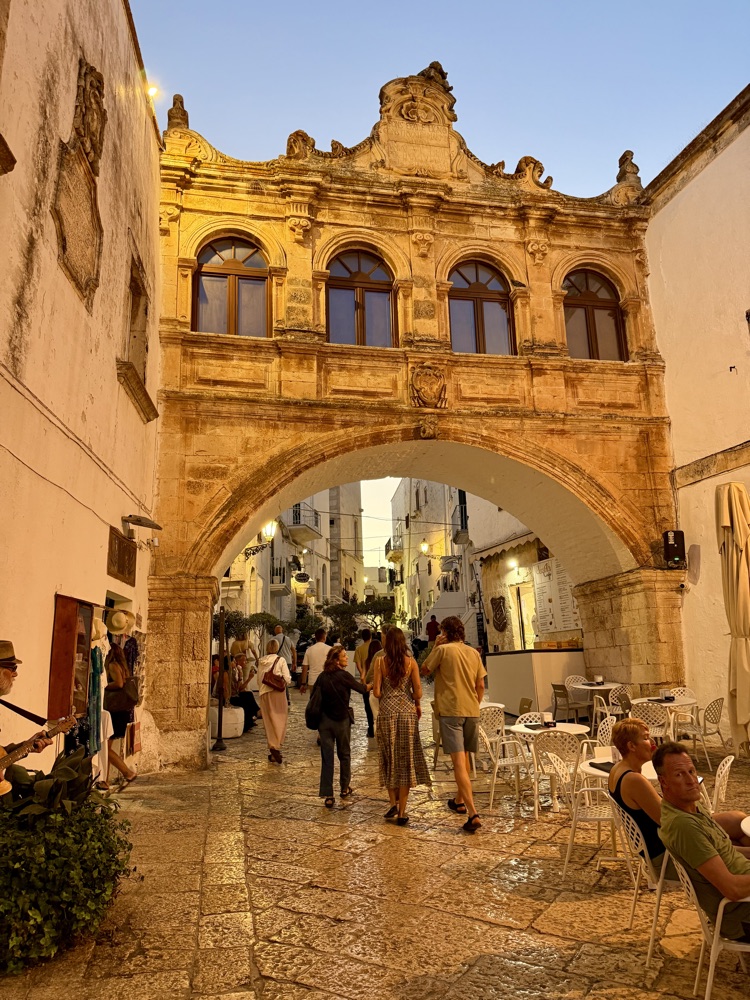 Verzierter Steinbogen über einer Gasse in Ostuni, darunter Menschen an Restauranttischen